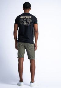 Camiseta negra con un gráfico dorado que presenta la palabra "PETROL" y una escena de playa, combinada con pantalones cortos verde oliva, con los dobladillos enrollados, y zapatillas blancas.