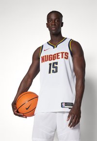 Biela basketbalová dres s nápisom "NUGGETS" v červenej a žltej farbe, s číslom "15". Model drží oranžovú basketbalovú loptu Nike. Biele šortky s textúrovaným vzorom.