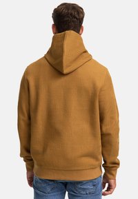 Sweat à capuche de couleur moutarde avec un motif texturé, présentant un ourlet et des poignets côtelés. Vu de dos, mettant en avant le détail de la capuche.