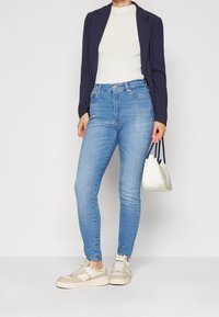Jeans ajustados azules, blusa blanca texturizada, chaqueta oscura y zapatillas crema. El conjunto incluye un pequeño bolso blanco con correas negras.