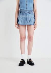 Denim miniszoknya plisszírozott dizájnnal, világoskék színben, övhurkokkal. Fekete lóferek és fehér bokazokni kíséretében.