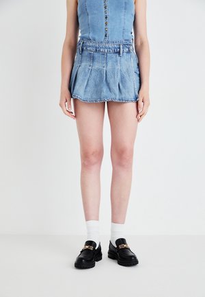 Denim mini-jurk met geplooid ontwerp, lichtblauwe kleur en riemlussen. Gecombineerd met zwarte instappers en witte enkelsokken.
