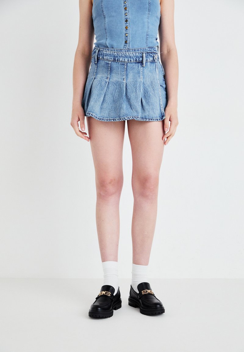 Denim miniszoknya plisszírozott dizájnnal, világoskék színben, övhurkokkal. Fekete lóferek és fehér bokazokni kíséretében.