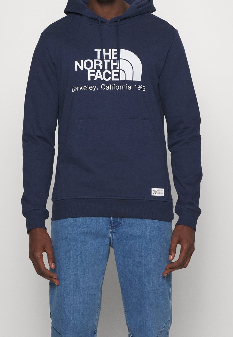Personne portant un sweat à capuche bleu marine avec le texte "The North Face Berkeley, California 1966" et un jean bleu sur un fond uni.