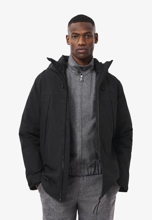 Zwarte waterdichte jas met een capuchon, ritssluiting aan de voorkant en zijzakken, gelaagd over een grijze fleece zip-up top met een hoge kraag.