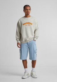 Lys grå hettegenser med oransje og grønn "homeboy" grafikk, kombinert med blå cargo-shorts og hvite joggesko med gule detaljer.