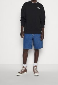 Musta collegepaita logolla, siniset farkkushortsit ja ruskeat korkeavartiset tennarit. Ominaisuuksiin kuuluu resorit hihansuuissa ja helmassa sekä näkyvät saumat.