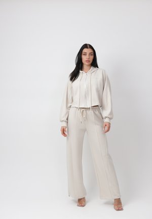 Frau steht vor weißem Hintergrund, trägt einen cremefarbenen Kapuzenpullover mit Reißverschluss und passende weite Hosen sowie transparente Sandaletten mit hohem Absatz.