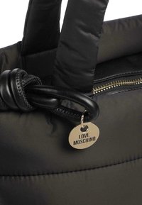 Bolsa de tote negra de nailon con asas acolchadas, cremallera dorada y etiqueta circular adjunta grabada con "LOVE MOSCHINO". Textura suave, diseño minimalista.