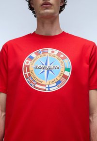 Rød bomulds T-shirt med et cirkulært grafisk motiv, der har en blå stjerne, tekst og forskellige nationalflag i et farverigt design.