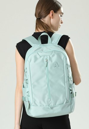 Vrouw draagt een mouwloos zwart topje en een lichtblauwe rugzak met een voorvak met rits en gewatteerde schouderbanden, met de rug naar de camera gekeerd.