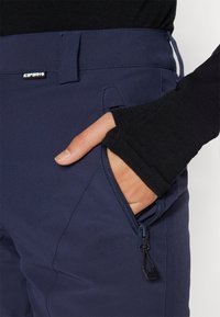 Mørkeblå stretchy bukser med en glidelås-lomme og en glatt overflate. Svart langermet skjorte synlig ved håndleddet, med ribbet tekstur.