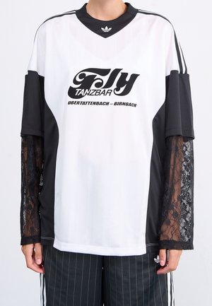 Person iført en hvid og sort Adidas-t-shirt med teksten "Fly Tanzbar Obertattenbach-Birnbach" og sorte blondeærmer under korte ærmer.
