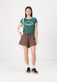 Camiseta verde corta con detalles y texto en blanco, pantalones cortos marrones con cordón y zapatillas negras con suela beige. Pequeño bolso blanco de hombro.