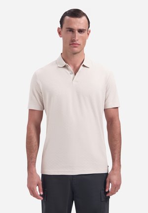 Man draagt een lichtbeige poloshirt met korte mouwen en donkere cargobroek, staand tegen een effen witte achtergrond.