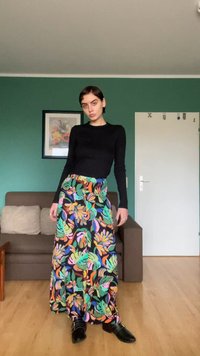 Μακριά, πολύχρωμη φούστα με μοτίβο τροπικών φύλλων σε πράσινο, πορτοκαλί και ροζ, συνδυασμένη με στενή μακρυμάνικη μαύρη μπλούζα και μαύρα παπούτσια.