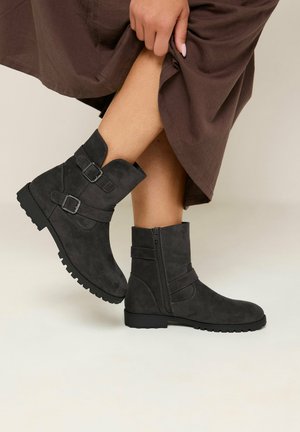 Mujer con botines de ante negro con hebillas y cremallera, levantando una falda marrón oscuro para mostrar las botas.
