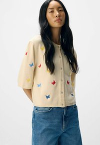 Cardigan crème à manches courtes, orné de papillons brodés multicolores en rouge, bleu et jaune. Fini avec un devant à boutons.