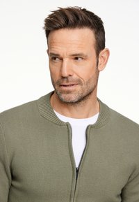 Maglione verde oliva con zip, colletto a coste, tessuto a maglia testurizzato, indossato sopra una camicia bianca, con chiusura lampo liscia.
