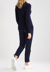 Kobieta odchodząca, ubrana w granatową bluzkę z długim rękawem, pasujące spodnie z rozcięciami na kostkach i białe trampki na jednolitym tle.