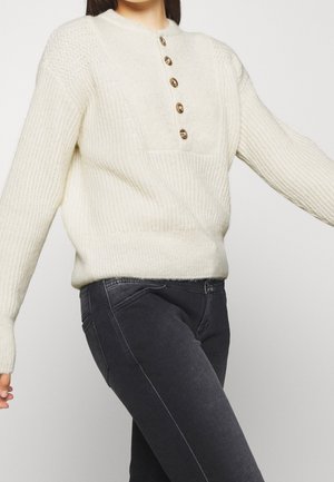 Femme portant un pull en maille crème avec cinq boutons en bois et un jean slim gris foncé sur un fond uni.
