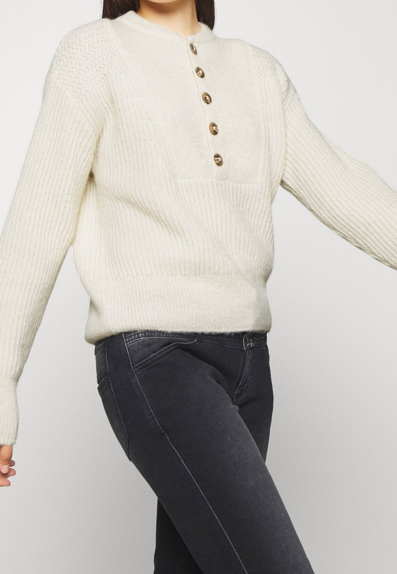 Femme portant un pull en maille crème avec cinq boutons en bois et un jean slim gris foncé sur un fond uni.