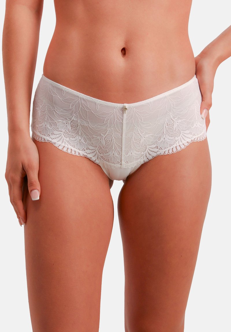 Białe koronkowe bokserki z falistymi brzegami, ozdobione delikatnymi kwiatowymi wzorami i centralnym szwem. Pas jest gładki, z minimalną ilością metalowych elementów.