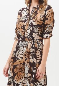 Donna che indossa un vestito button-up con stampa tropicale a foglie marrone, beige e bianco, con cintura in vita e maniche arrotolate.