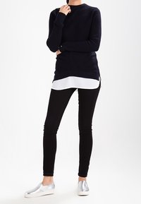 Marinblå stickad tröja med rund halsringning, lager på lager över en vit skjorta, kombinerad med svarta tajta jeans och silverfärgade slip-on skor.