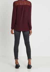 Blusa bordeaux con pannello superiore in pizzo, maniche lunghe e vestibilità ampia, abbinata a jeans grigi attillati e scarpe slip-on nere.