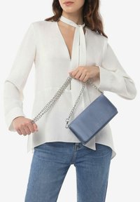 Borsa a tracolla in pelle blu chiaro con una cinghia a catena, caratterizzata da una forma rettangolare e una texture liscia. Abbinata a una blusa bianca e jeans.