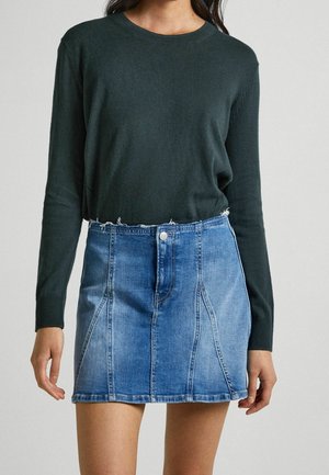 Kvinde iført en mørkegrøn langærmet sweater stoppet ind i en lyseblå jeans mini nederdel med rå kant.
