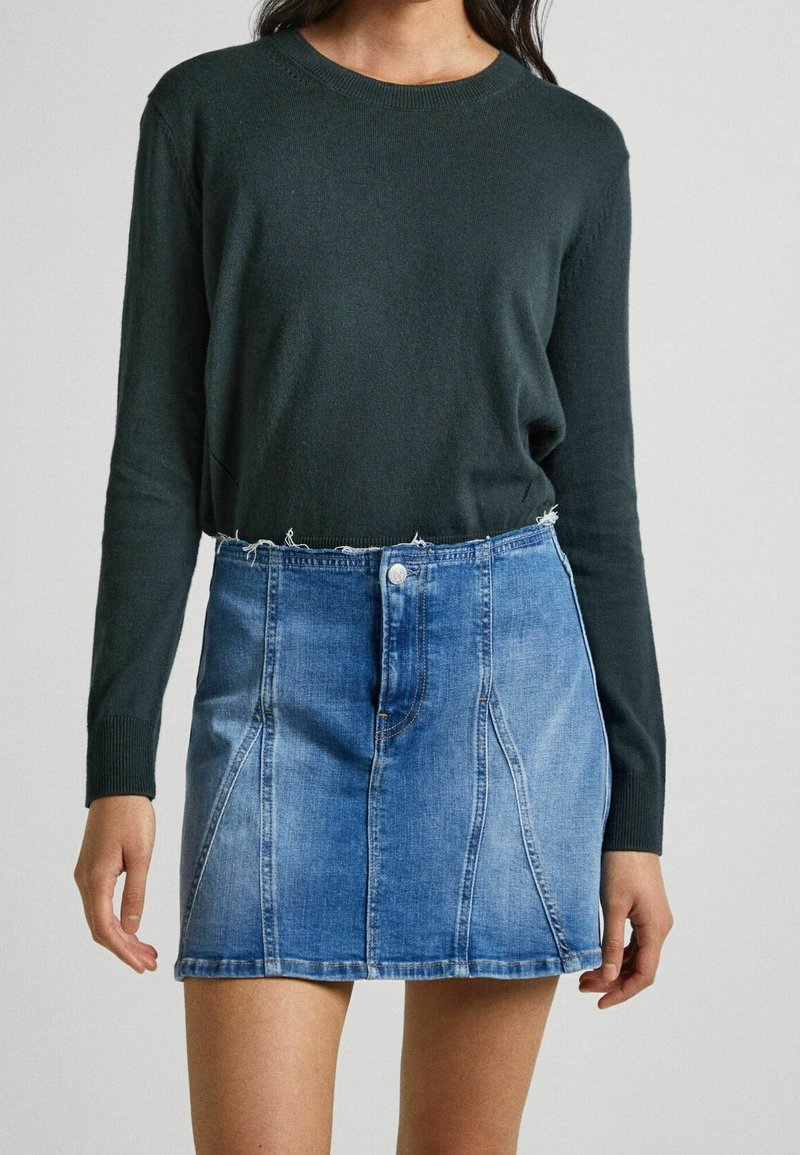 Femme portant un pull vert foncé à manches longues rentré dans une mini-jupe en jean bleu clair avec un ourlet brut.