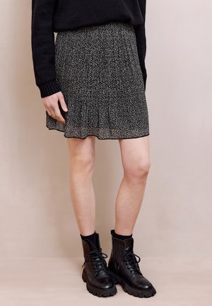 Osoba ubrana w czarną spódnicę w wzory, czarny sweter oraz czarne sznurowane botki wojskowe, stojąca na tle jasnobeżowego tła.