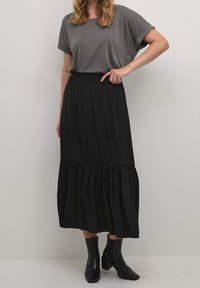 T-shirt grigio a maniche corte abbinato a una gonna lunga nera a strati con vita smock. Stivaletti neri completano l'outfit.