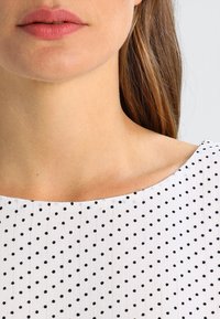 Top in tessuto bianco con motivo a pois neri, caratterizzato da un ampio scollo a barca e una superficie testurizzata, che mette in risalto un collo liscio.