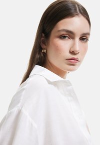 Hvid button-up skjorte med et struktureret stof, der har en krave og løst fit. Gyldne geometriske øreringe tilføjer accentdetaljer. Neutral baggrund.