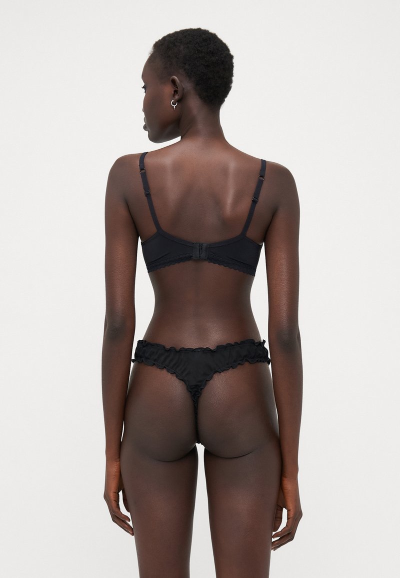 Reggiseno in pizzo nero abbinato a perizoma con bordi arricciati, dotato di spalline regolabili e chiusura a gancio, che mostra una superficie liscia.