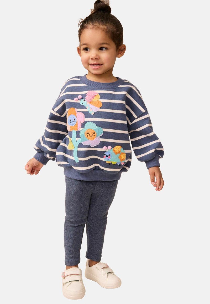 Gestreifter blauer Sweatshirt mit buntem Applikationsblumen und Schmetterlingen, kombiniert mit marineblauen Leggings und weißen Turnschuhen mit pastellfarbenen Akzenten.