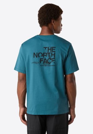 Mand iført en teal kortærmet T-shirt med sort "The North Face" logo og bjerggrafik på ryggen, sammen med sorte shorts.