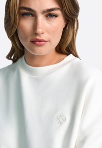 Balts sporta krekls ar ribotiem apkakles malām un izšūtu logotipu. Mīksta tekstūra un brīvs siluets.