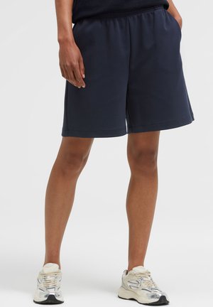 Adult standing wearing black knee-length shorts, black top, and white athletic sneakers with laces on a light background.