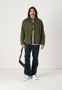 Veste en coton vert avec poche poitrine, boutons noirs, portée sur un T-shirt blanc, associée à un pantalon bleu marine et des baskets blanches. Sac à bandoulière noir.
