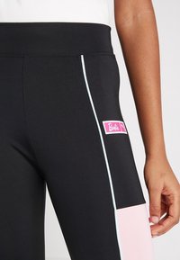 Zwarte sportleggings met een roze paneel, lichtblauwe biezen aan de zijkanten en een rechthoekig wit logo met roze letters. Gladde textuur.