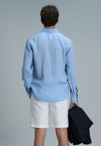 Helles blaues Hemd mit langen Ärmeln aus Leinen, kombiniert mit weißen Shorts. Das Hemd hat einen Knopfkragen und einen abgerundeten Saum. Hält eine dunkle Jacke.