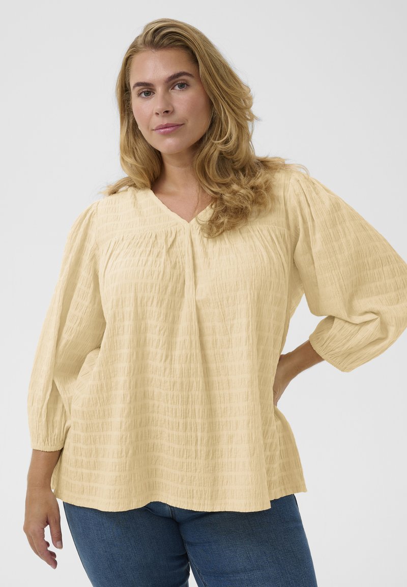 Femme aux longs cheveux blonds portant une blouse ample et texturée jaune pâle à manches trois-quarts et un jean bleu, posant devant un fond blanc.