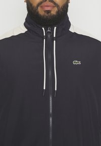 Marinblå vindjacka med hög krage, crèmefärgade axelaccenter och vita dragsnören. Har en framzipper och broderad logotyp på bröstet.