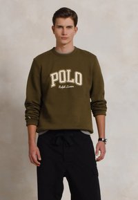 Mladic u maslinastozelenoj Polo Ralph Lauren dukserici i crnim hlačama, koji stoji uz jednoličnu neutralnu pozadinu.