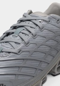 Gros plan sur une chaussure de sport grise avec une maille tissée, des superpositions structurées et des lacets épais en forme de croix noués en haut.