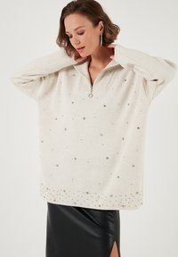 Beżowy sweter na zamek z prążkowanym kołnierzem i ozdobnymi srebrnymi detalami, zestawiony z czarną skórzaną spódnicą.